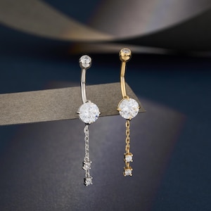 May include: Two belly button rings, one silver-toned and one gold-toned, each featuring a large, clear, round gemstone. The silver ring has a chain with three smaller gemstones, while the gold ring has a similar chain. Both are curved.