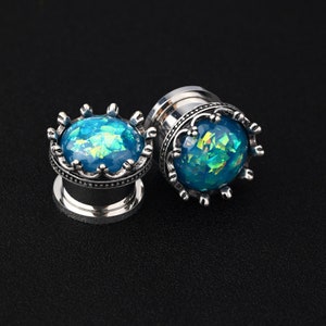 Könnte beinhalten: Zwei silberfarbene Ohr-Dehnstäbe mit Kronen-Design. Jeder Dehnstab hat eine blau-grüne, schillernde Kugel, die in einer detaillierten, kronenförmigen Lünette gefasst ist. Die Dehnstäbe werden vor einem schwarzen Hintergrund gezeigt.