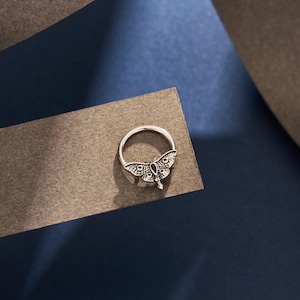 May include: A silver ring featuring a detailed moth design. The moth has intricate wing patterns and a small, dark stone at its center. The ring is set against a dark blue and gray background, highlighting the ring's metallic sheen.