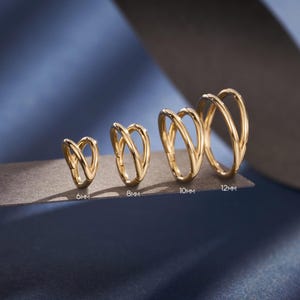以下が含まれることがあります： 6mmから12mmまでの、さまざまなサイズの4つのゴールドカラーのイヤーカフ。各カフは、互いに交差するダブルバンドデザインが特徴です。ジュエリーは、青い背景に対して暗い表面に表示されています。