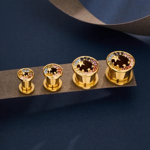 Puede incluir: Cuatro expansores de oreja dorados con diseño de estrella. Cada expansor está adornado con un anillo de piedras preciosas de colores. El centro de cada expansor es marrón oscuro. Los expansores se muestran sobre una superficie gris oscuro sobre un fondo azul oscuro.