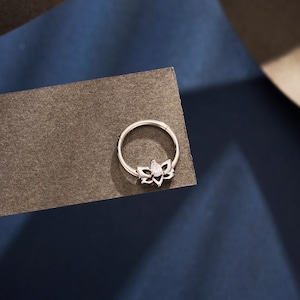 May include: A silver nose ring with a lotus flower design and a clear gemstone centre. The ring is circular with a hinged closure. The lotus flower has multiple detailed petals. The ring is made of silver.