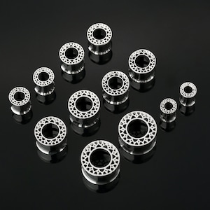 Puede incluir: Una colección de expansores de oreja plateados con un diseño calado decorativo. Los expansores son cilíndricos con un borde acampanado y un patrón geométrico. Están dispuestos sobre una superficie negra reflectante.