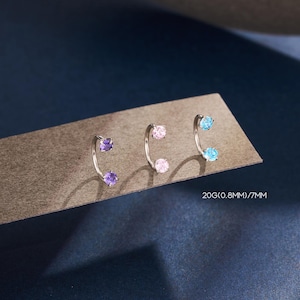May include: Three silver-coloured curved earrings, each with a gemstone at either end. The gemstones are purple, pink, and light blue. The text "20G(0.8mm)/7mm" is visible.