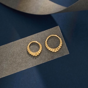 Könnte beinhalten: Zwei goldfarbene Creolen mit einem strukturierten, sonnenstrahlenartigen Design. Die Ohrringe werden auf einer grauen Oberfläche vor einem dunkelblauen Hintergrund präsentiert. Die Ohrringe sind klein und scheinen aus Metall zu sein.