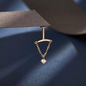 May include: A gold-colored earring with a small diamond pendant. The earring has a post that connects to a horizontal bar, from which a chain descends to hold the diamond. The design is simple and elegant, with a focus on the gemstone.