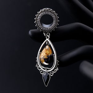 Puede incluir: Túnel de oreja plateado con centro transparente y anillo exterior decorativo. Un colgante en forma de lágrima cuelga debajo, con pan de oro encapsulado en resina y una piedra preciosa negra. La joya se muestra sobre un fondo negro.