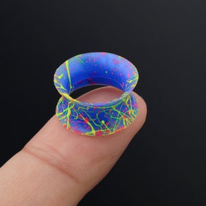May include: A blue silicone ear gauge with a splatter paint design in yellow, pink, and green.