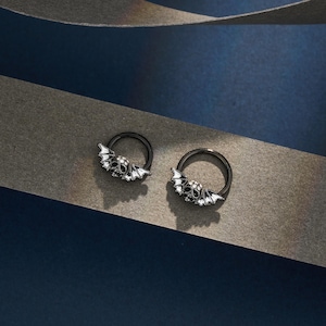 Puede incluir: Dos pendientes de aro negros con un diseño de calavera y alas de murciélago. Las calaveras tienen detalles blancos, y las alas también son blancas. Los pendientes se muestran sobre un fondo azul oscuro y gris.