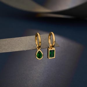 May include: A pair of gold hoop earrings with emerald green gemstones. One earring has a teardrop-shaped stone, the other a rectangular stone. The earrings are set against a dark blue backdrop.