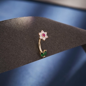May include: A gold-colored belly button ring with a floral design. The flower has white and red gemstones, and the bottom features two green leaf-shaped gems. The jewelry is curved and set against a dark background.