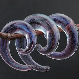 May include: Two clear glass spiral ear gauges with a purple and blue glitter finish.