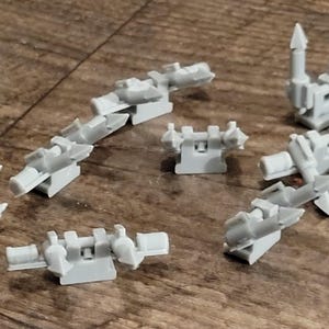 May include: A collection of light gray, 3D-printed miniature components, possibly for a model or game. The pieces vary in shape, including spiky, angular designs and rectangular blocks. The objects are scattered on a wooden surface.