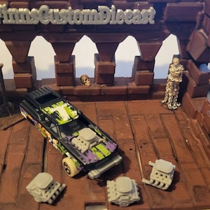 May include: A black and green toy car with a white skull and crossbones design. The car is on a brown and gray playset with a guillotine, a skull, and a skeleton figure. The playset has the words "Films Custom Diecast" on the top.