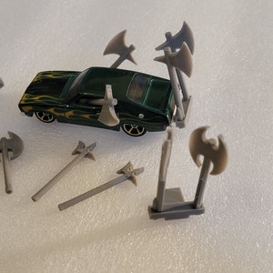 May include: A green toy car with yellow and green flames, surrounded by several gray miniature axes. Some axes are mounted on gray stands. The scene is set on a white surface.