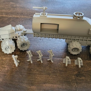 May include: A gray, 3D printed model of a futuristic, military-style vehicle with a large tank trailer. The vehicle has six wheels and a gun turret on top. The trailer has a large tank and a small window. The model is on a wooden surface.