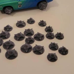 May include: A collection of small, dark gray, spiky objects, resembling miniature spiked wheels, are scattered on a light surface. A teal and blue toy car with matching spiked wheels is visible in the background, suggesting a theme of automotive accessories or toys.