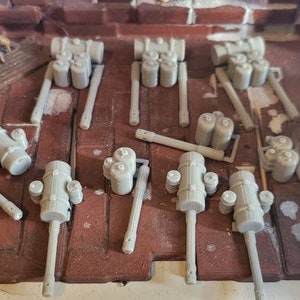 May include: A collection of gray plastic model cannons with barrels and wheels. The cannons are arranged in a grid pattern on a brown surface.