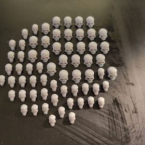 May include: A large collection of small, gray, plastic skulls arranged in a circular pattern on a dark gray surface.