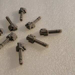 May include: Eight gray plastic miniature cannons arranged in a circle on a white textured surface.