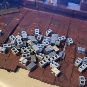 May include: A pile of gray concrete blocks, scattered on a brown surface. The blocks are small and have a rough texture.