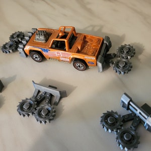 May include: Orange toy truck with a black and white American flag design and multiple gray, gear-like attachments. The truck is a Hot Wheels toy.