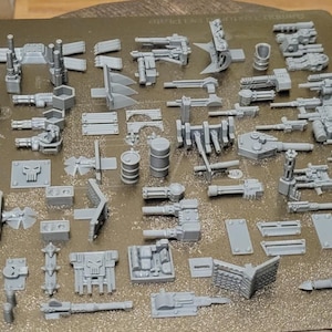 May include: A collection of unpainted, gray plastic miniature war game components. The assortment includes various weapons, barrels, and structural elements, ideal for tabletop gaming enthusiasts. The pieces are arranged on a brown surface.