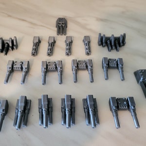 May include: A collection of 19 grey 3D printed miniature cannons, some with double barrels and some with triple barrels. The cannons are arranged in a triangular pattern on a light grey surface.