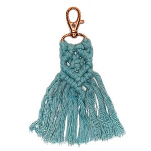 May include: A teal blue macrame keychain with a copper-colored metal clasp. The keychain has a fringe of tassels.