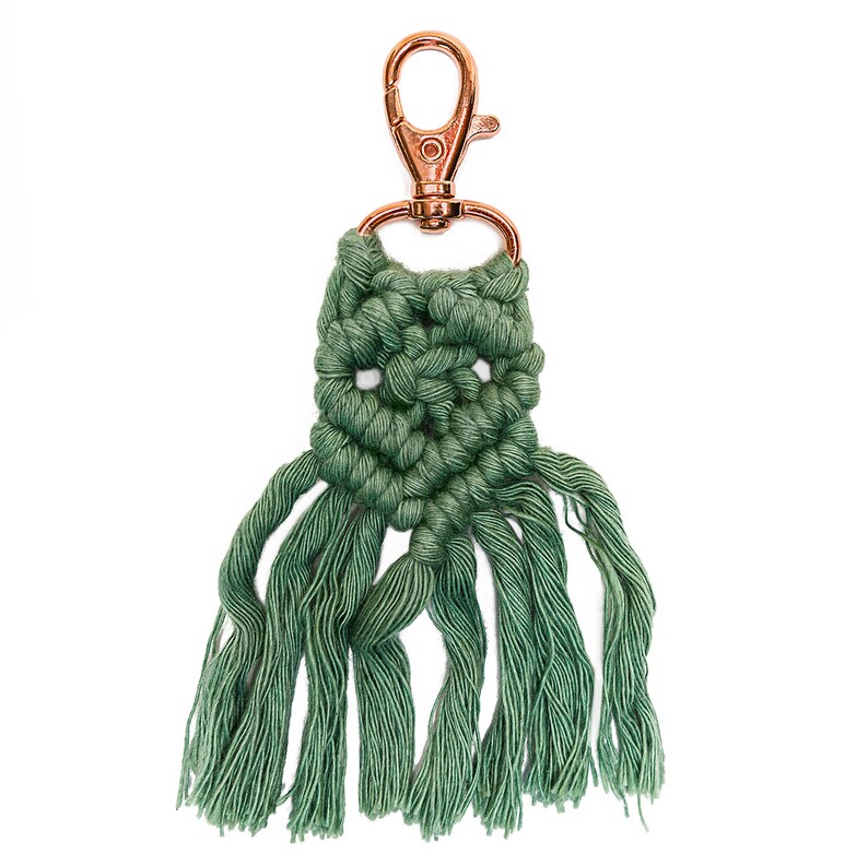 May include: A green macrame keychain with a copper-colored metal clasp. The keychain has a fringe of tassels.