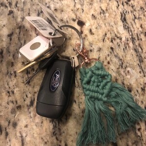 May include: A set of keys with a green tassel keychain. The keychain is made of macrame and has a fringe. The keys include a black Ford car key and several other keys.