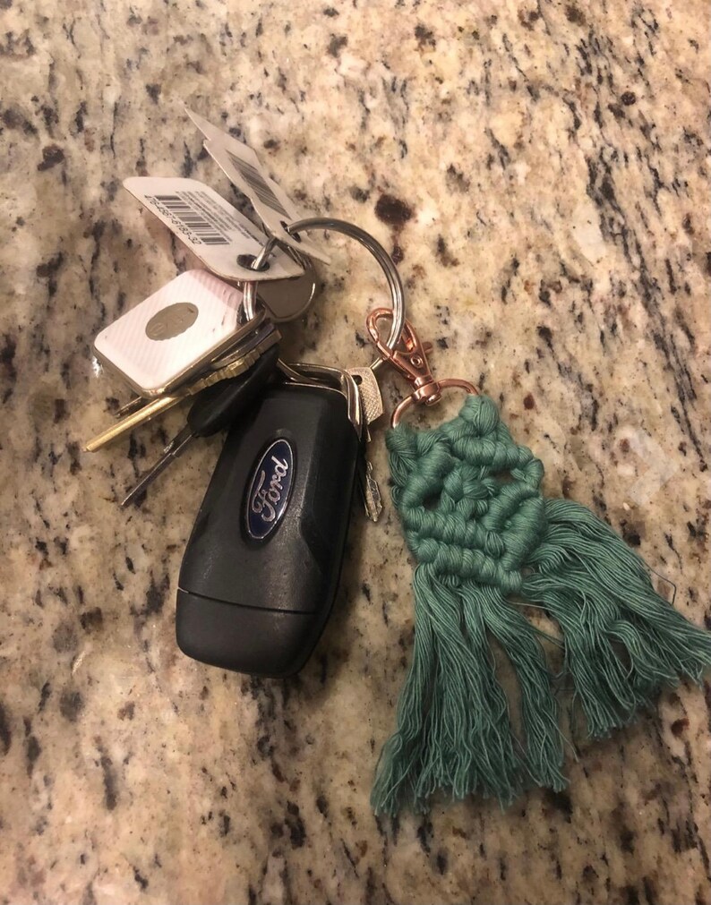 May include: A set of keys with a green tassel keychain. The keychain is made of macrame and has a fringe. The keys include a black Ford car key and several other keys.