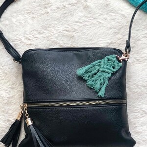 May include: A black leather crossbody bag with a zipper closure and two black tassels. The bag has a teal blue tassel charm attached to the zipper pull.