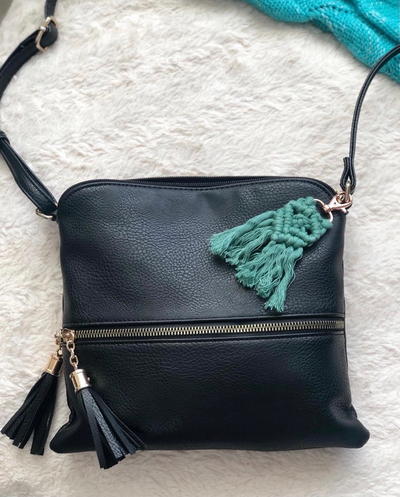 May include: A black leather crossbody bag with a zipper closure and two black tassels. The bag has a teal blue tassel charm attached to the zipper pull.