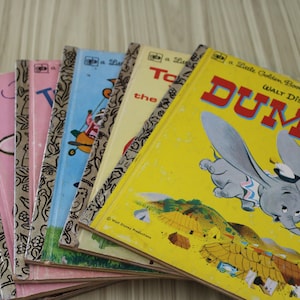 May include: A collection of vintage children's books, including a yellow "Dumbo" book with an illustration of the elephant. Other books have pink, blue, and yellow covers. The books are arranged in a fan shape.