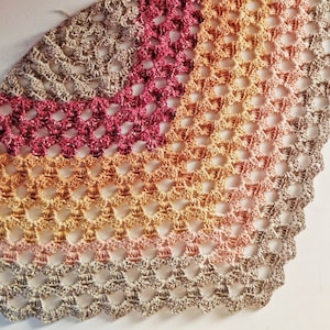 May include: A crocheted shawl with a gradient of colors, starting with a light beige, then transitioning to peach, orange, and finally a deep burgundy. The shawl is made with a delicate openwork stitch pattern.