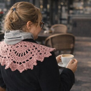 May include: A light pink, crocheted collar with a scalloped edge. The collar is worn over a black jacket and a gray hooded sweatshirt. The person is holding a white cup of coffee.