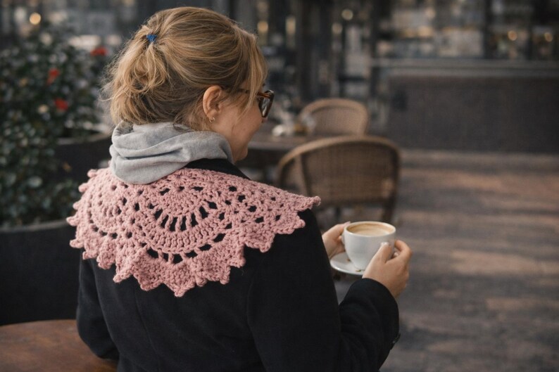 May include: A light pink, crocheted collar with a scalloped edge. The collar is worn over a black jacket and a gray hooded sweatshirt. The person is holding a white cup of coffee.