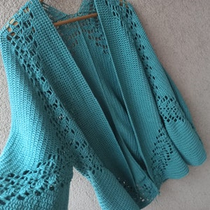 Puede incluir: Un cárdigan de color azul turquesa tejido a crochet con un patrón de encaje. El cárdigan tiene un cuello en V profundo y mangas largas.