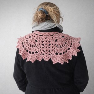 May include: A light pink, crocheted shawl with a scalloped edge. The shawl is circular in shape and features an open, lacy design. The person is wearing a black coat and a gray hooded sweatshirt.