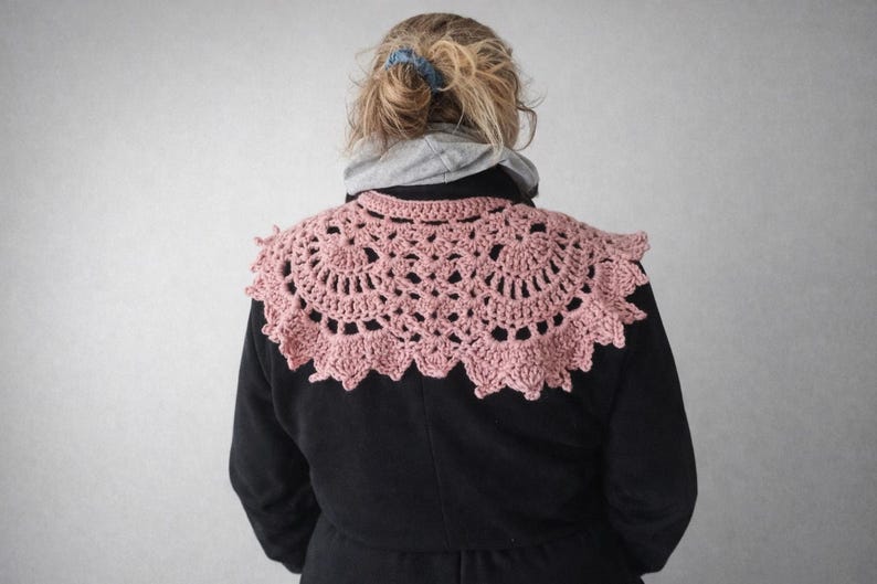 May include: A light pink, crocheted shawl with a scalloped edge. The shawl is circular in shape and features an open, lacy design. The person is wearing a black coat and a gray hooded sweatshirt.