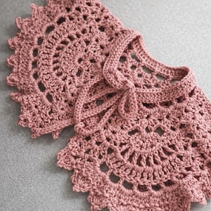 Könnte beinhalten: Ein staubrosa gehäkelter Kragen mit gewelltem Rand und Bindeband. Das Lochmuster weist kreisförmige Muster und ein strukturiertes Aussehen auf. Dieses handgefertigte Accessoire verleiht einen Hauch von Vintage-Charme.