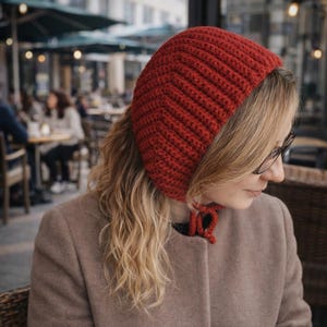 May include: A red crocheted hat with a rounded shape and a tie closure. The hat is made of a textured yarn and is worn by a person with blonde hair and glasses. The person is wearing a brown coat.