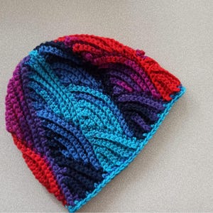 May include: A handmade, colorful crochet beanie hat. The hat features a textured design with a gradient of colors, including red, purple, blue, and turquoise. The hat is made of soft yarn and is suitable for cold weather.