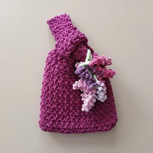 May include: A deep magenta crochet handbag featuring a thick, textured stitch pattern and a single integrated handle. Attached to the front is a decorative cluster of crocheted flowers in shades of light pink, lavender, and purple with green stems.