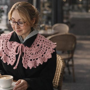 May include: A light pink, crocheted collar with a scalloped edge and tie closure. The collar is worn over a black coat. The image shows a woman wearing glasses and holding a cup of coffee at an outdoor cafe.