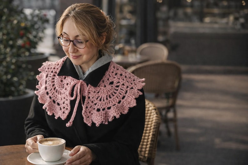 May include: A light pink, crocheted collar with a scalloped edge and tie closure. The collar is worn over a black coat. The image shows a woman wearing glasses and holding a cup of coffee at an outdoor cafe.