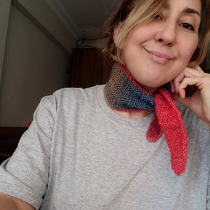 May include: A woman wearing a grey t-shirt with a multi-coloured crocheted scarf around her neck. The scarf is red, blue and brown.