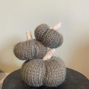 Op de afbeelding: Een stapel van drie gebreide pompoenen in verschillende maten. De pompoenen zijn grijsgroen van kleur met crèmekleurige stelen. De pompoenen zijn geplaatst op een donker, rond oppervlak, wat een herfstthema suggereert.