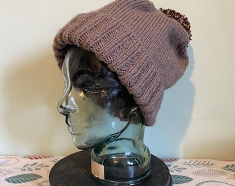Puede incluir: Un gorro de punto marrón con un patrón texturizado y un pequeño pompón en la parte superior. El sombrero se exhibe en una cabeza de maniquí de cristal. La cabeza del maniquí está sobre un soporte circular negro, con un mantel estampado en el fondo.
